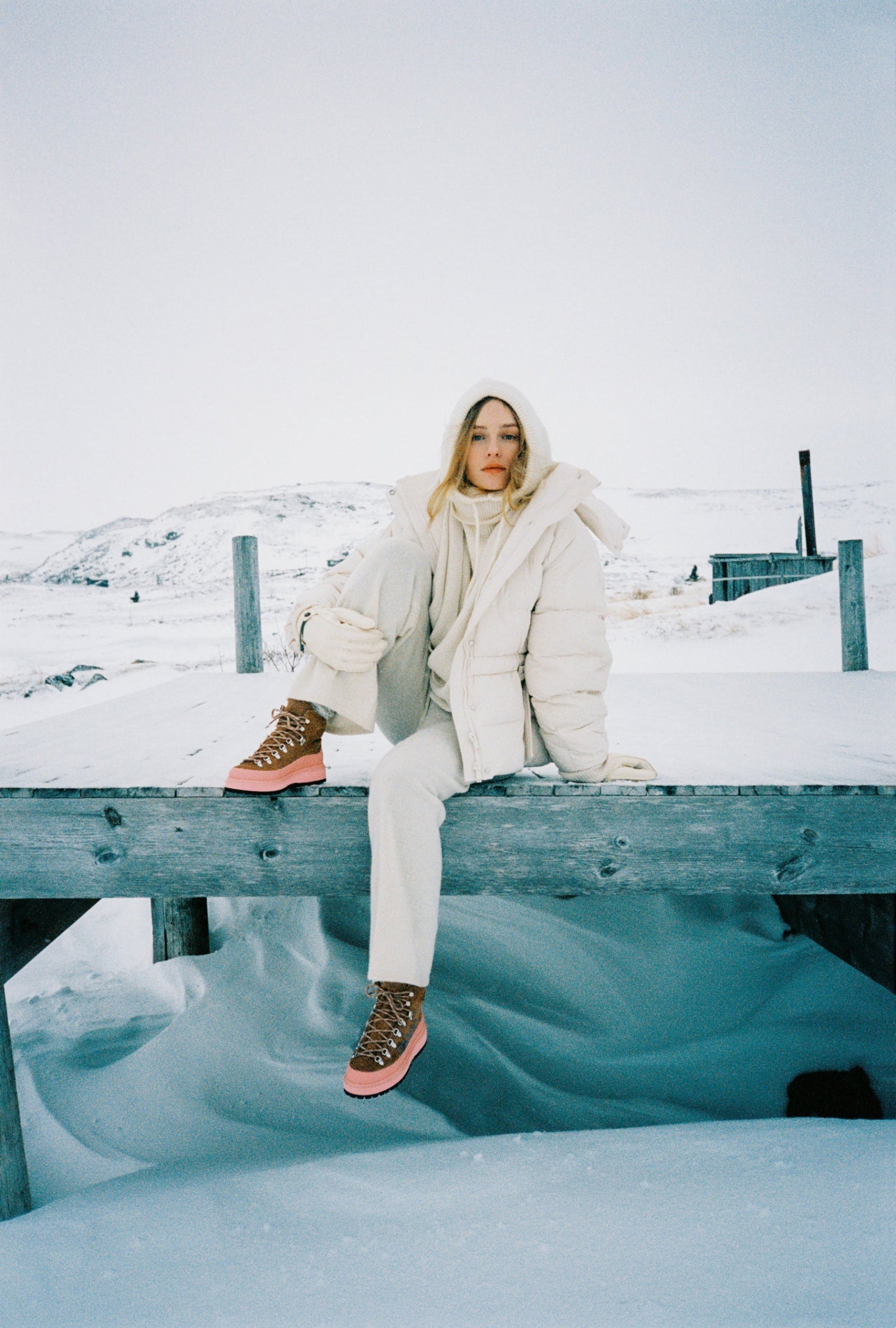 Person sitting on a snowy dock wearing a white coat and pink winter shoes.