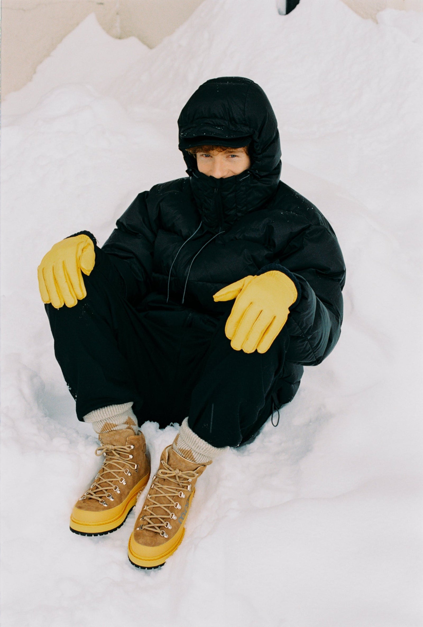 Person wearing a black snowsuit with yellow gloves and boots sitting in the snow with colorful winterboots